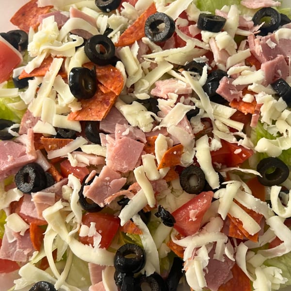 Close-up of a Ciceros Pizza salad with shredded cheese, pepperoni, ham, black olives, and tomatoes.